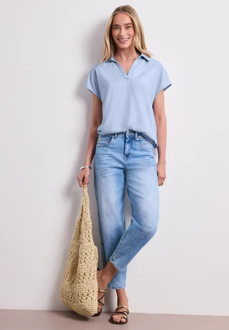 Frau in hellblauem Poloshirt, lässigen Jeans, Sandalen und großem gehäkeltem Strandbeutel.