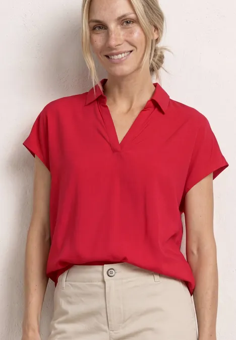 Vrouw in een rood T-shirt met korte mouwen en een beige broek voor een lichte achtergrond.