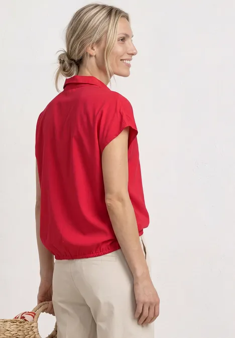 Vrouw in een rode blouse met korte mouwen en beige broek draagt een kleine strooien tas.
