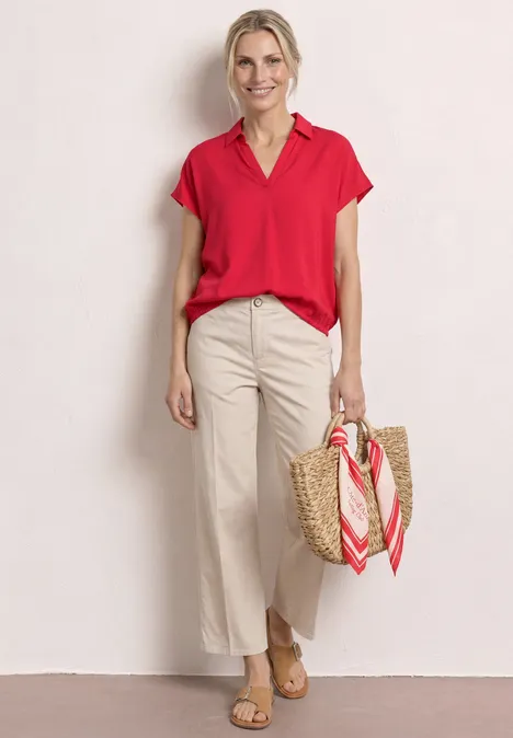 Vrouw in rode blouse en witte broek, met een rieten tas met sjaal tegen een lichte achtergrond.