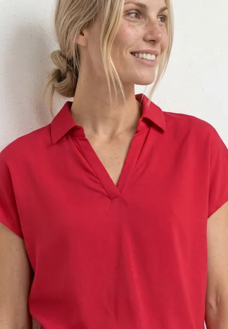 Vrouw voor witte muur met rode top met korte mouwen en V-hals.