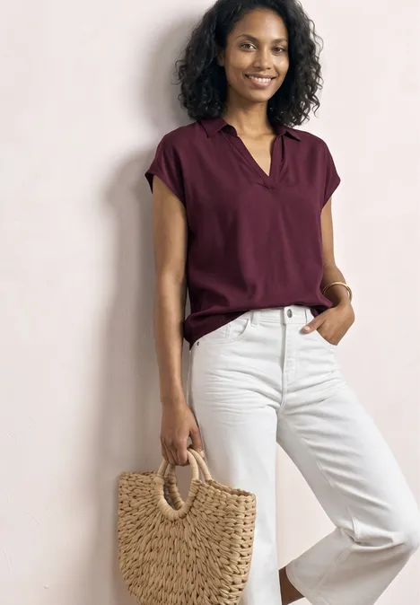 Een vrouw draagt een bordeaux top, witte spijkerbroek en houdt een strooien tas met ronde handvatten.