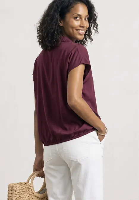 Een vrouw staat zijwaarts, draagt een los bordeauxrood shirt, witte broek en houdt een rieten tas.