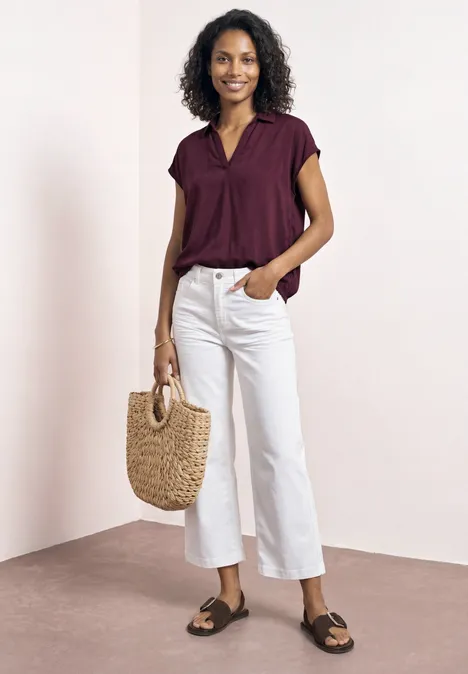 Vrouw in bordeaux blouse, witte wijde cropped broek, zwarte slippers en strooien tas voor neutrale achtergrond.