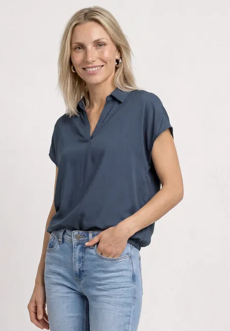 Persoon in donkerblauw kort overhemd en lichte jeans, ontspannen staand voor een neutrale achtergrond.