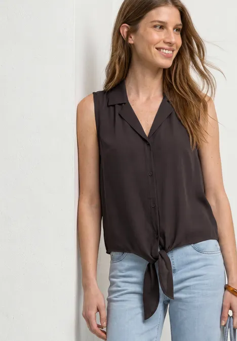 Eine junge Frau trägt eine ärmellose, knöpfbare Bluse mit Knoten vorne und eine helle Jeans.