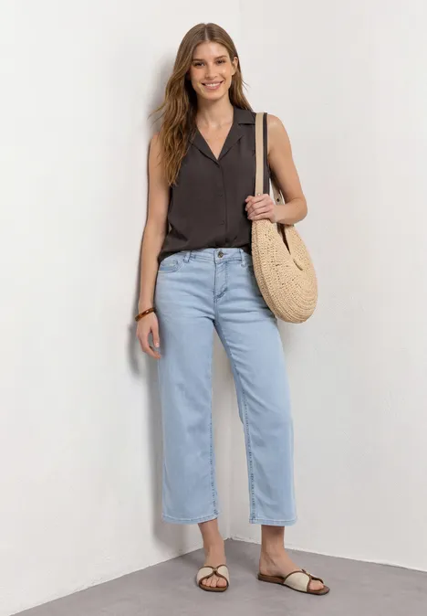Frau in dunklem ärmellosem Top, hellblauen Jeans, braunen Sandalen und Strohtasche vor weißem Hintergrund.