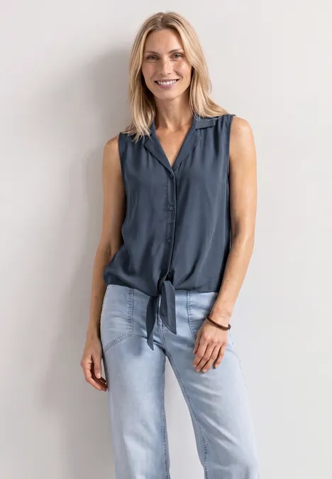 Eine blonde Frau lehnt vor weißer Wand, trägt ärmellose dunkle Bluse mit Knoten und helle Jeans.