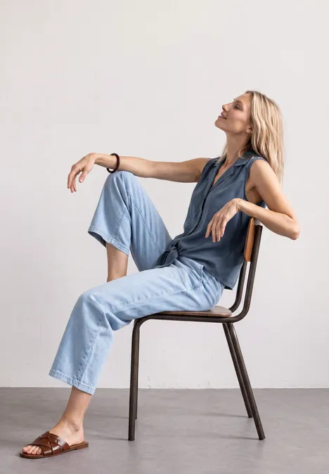 Frau sitzt entspannt seitlich auf Stuhl, trägt hellblaue Denim-Hose, mit erhobenem Blick, minimalistischer Hintergrund.