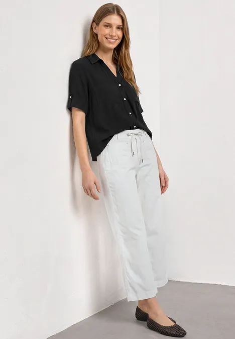 Chemisier à manches courtes à revers Black Femme appuyée contre un mur blanc, portant une blouse noire, un pantalon large blanc et des ballerines noires.