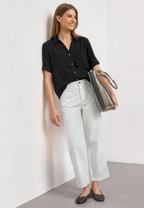 Chemisier à manches courtes à revers Black Une femme appuyée contre un mur, chemise noire, pantalon blanc, chaussures plates et grand sac à main.