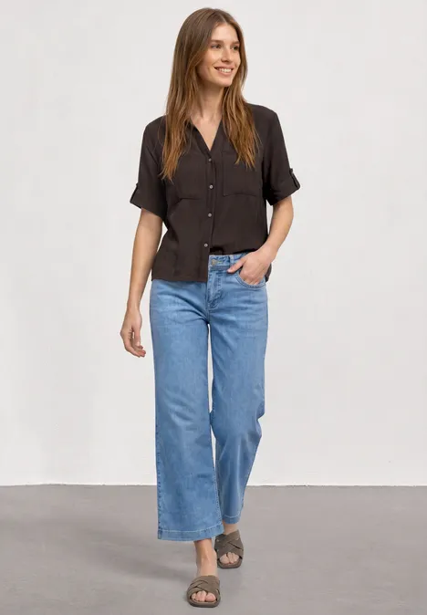 Frau in schwarzem kurzärmeligem Hemd, weiten blauen Jeans und Sandalen vor neutralem Hintergrund.