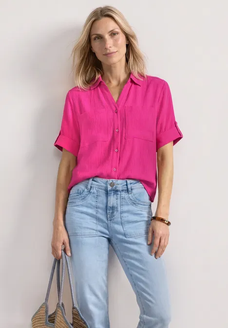 Chemisier à manches courtes à revers magenta dream Femme en chemise rose vif et jean clair, sac gris à la main, fond lumineux.