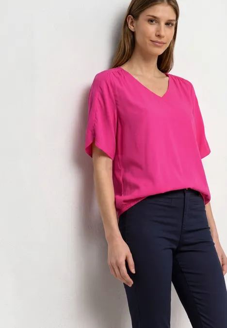 Frau lehnt entspannt an weißer Wand, trägt pinkes Shirt und schwarze Hosen.