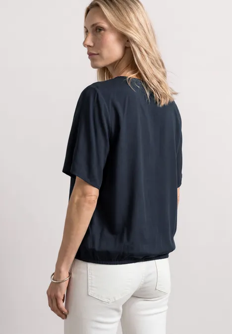 Frau mit blonden Haaren, schwarze Kurzarmbluse und weiße Jeans, Rückenansicht.