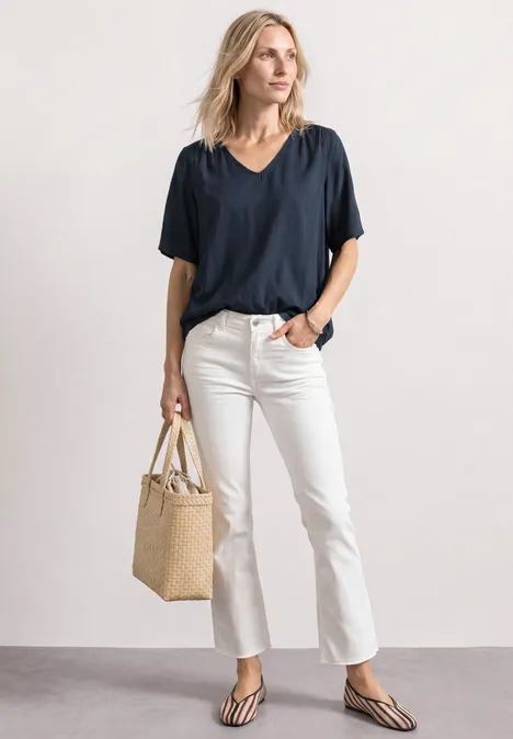 Frau in schwarzem V-Shirt, weißen Jeans und Sandalen, trägt einen geflochtenen Korbbeutel, steht vor weißem Hintergrund.