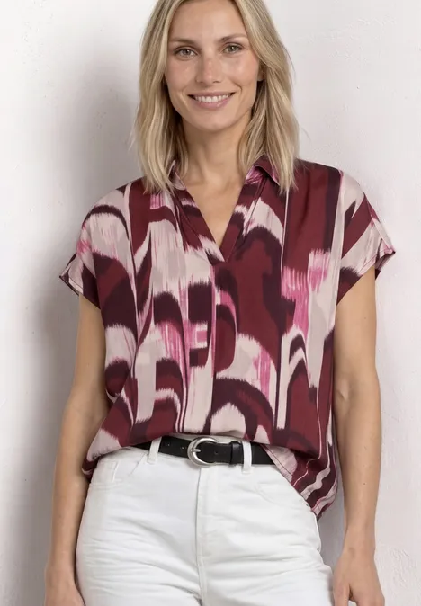 Vrouw in witte broek en bordeauxrood kort mouwig hemd met abstract patroon en zwarte riem.