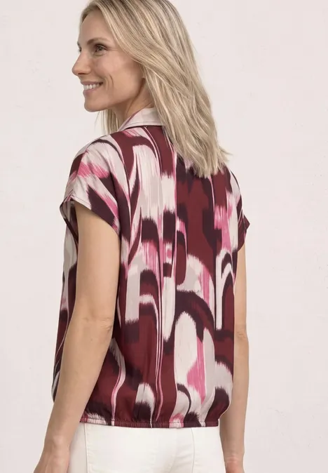 Een vrouw gezien van achteren met blond haar, draagt een blouse met korte mouwen in roze, wit en bordeaux met geometrisch patroon.