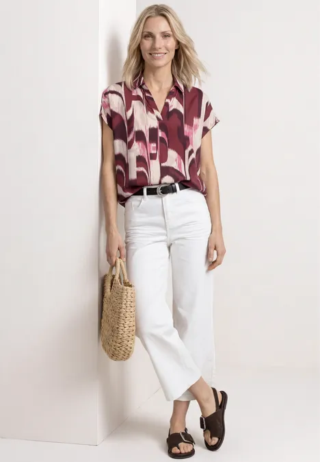 Vrouw tegen lichte achtergrond in kort shirt met print, witte jeans, bruine sandalen en rieten tas.