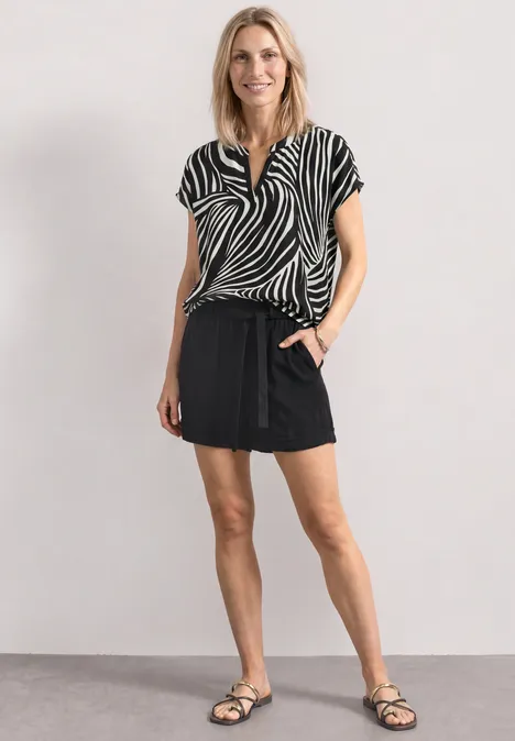 Blouse met split in de hals en luipaardprint Black Vrouw in zebrapatroon blouse, korte zwarte rok en sandalen tegen een neutrale achtergrond.