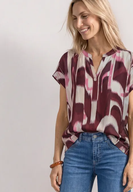 Blusenshirt mit Split Neck und Leo-Muster rustic rouge Frau trägt lockeres Kurzarmshirt mit abstraktem Rot-Weiß-Muster und Jeans.