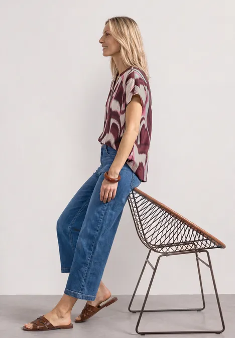Blusenshirt mit Split Neck und Leo-Muster rustic rouge Frau in gemusterter Bluse, weiten Jeans und braunen Sandalen, lehnt an einem Drahtstuhl.