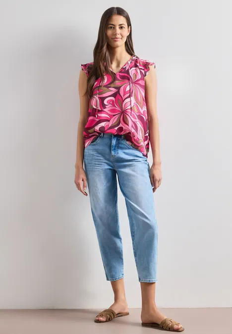 Frau in rosa geblümtem Top, hellblauen Jeans und flachen Sandalen vor neutraler Wand.