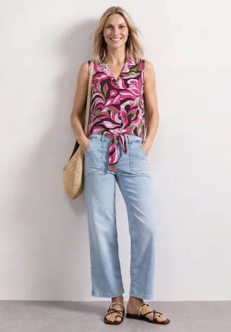 Frau in geblümter, pinker Bluse mit Knoten, blauen Jeans, Sandalen und Strohtasche vor weißem Hintergrund.