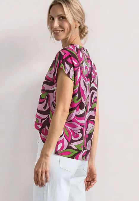 Frau trägt bunt gemustertes, kurzärmeliges Top mit abstraktem Swirl-Design und weiße Hose.