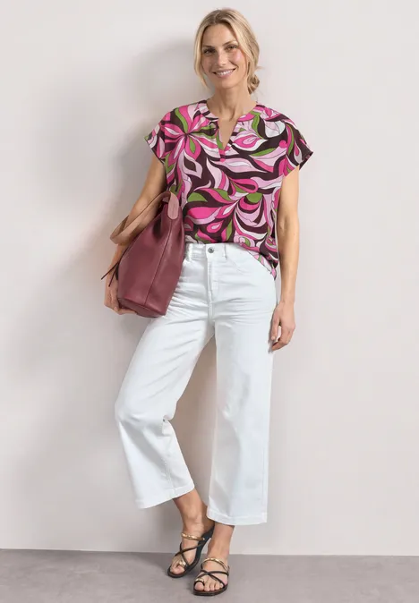 Frau mit hochgeknotetem Haar in pink gemusterter Bluse, weißer Culotte und schwarzen Sandalen, mit rosafarbenem Shopper.