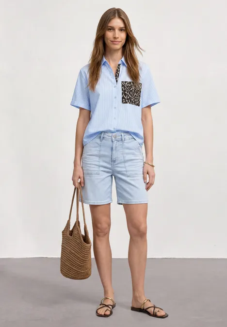Een vrouw draagt een lichtblauw overhemd met luipaardzak, jeansshort, zwarte sandalen en een rieten tas.