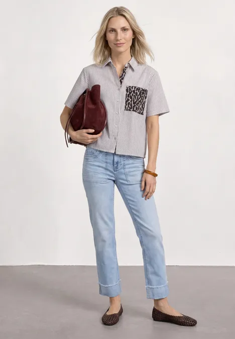 Frau trägt kurzärmliges, helles Hemd mit gemusteter Tasche, helle Jeans, flache Mokassins und trägt eine Handtasche.