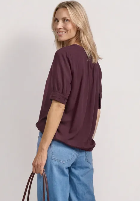 Eine blondhaarige Person von hinten mit einem purpurfarbenen Oberteil, blauen Jeans und Tasche.