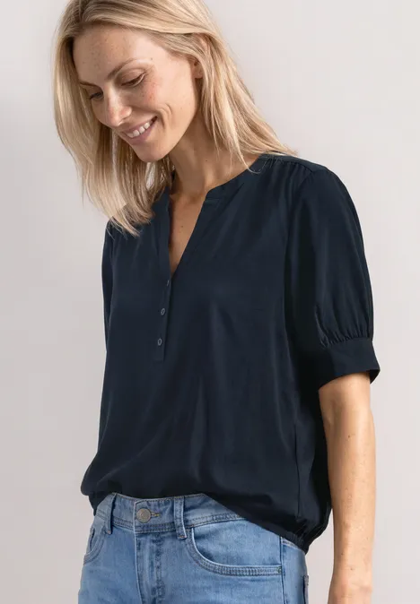 Frau in dunkelblauer Bluse und Jeans mit sanftem Lächeln vor hellgrauem Hintergrund.