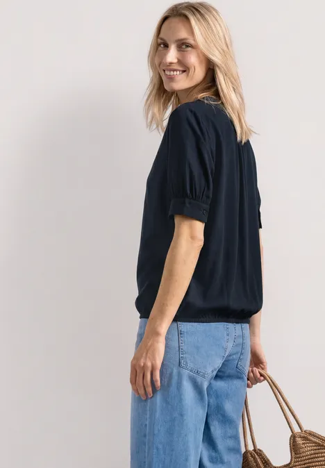 Seitliche Ansicht einer Frau in schwarzer Bluse, blauen Jeans und mit einer Strohtasche.