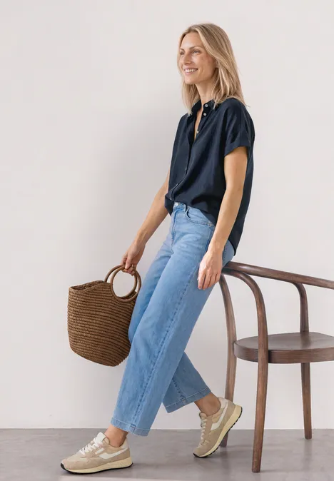 Frau im navy Hemd und hellen Jeans, trägt geflochtene Tasche, goldene Sneaker, steht neben Stuhl.
