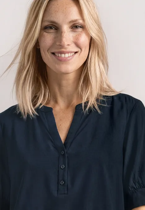 Eine Frau mit blonden, schulterlangen Haaren trägt eine dunkelblaue Bluse mit Knopfleiste, Gesicht unscharf.