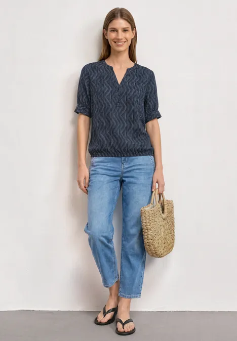Blouse met korte mouwen, gespleten hals en elastische zoom ashen blue Vrouw in T-shirt met patroon, blauwe jeans, slippers en rieten tas voor neutrale achtergrond.