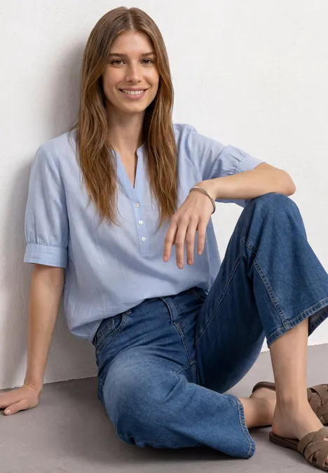 Une femme souriante assise décontractée en jean et chemise bleu clair devant un mur blanc.