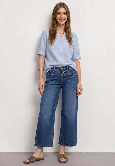 Une femme souriante en chemise bleu clair, jeans larges et sandales devant un fond blanc.