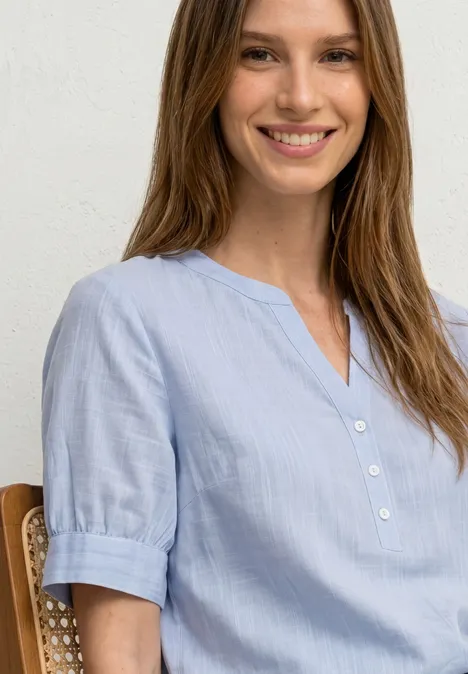 Jeune femme souriante assise sur une chaise, portant un chemisier bleu clair à manches courtes.