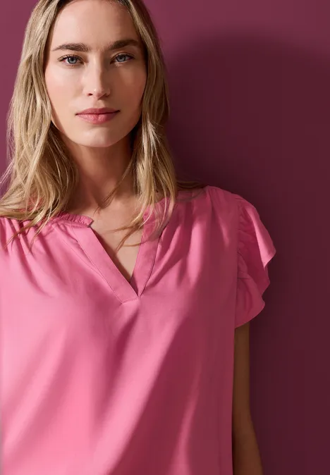 Eine blonde Frau in rosa Bluse vor dunklem rosafarbenen Hintergrund.