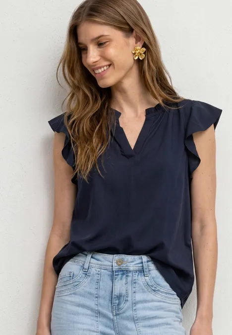 Lächelnde Frau trägt dunkelblaue Rüschenbluse und helle Jeans.