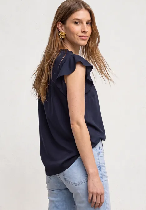 Eine lächelnde junge Frau in dunkelblauer Bluse mit Rüschenärmeln und Jeans seitlich fotografiert