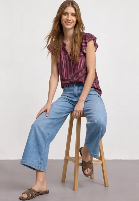 Eine Frau sitzt auf einem Hocker, trägt gestreiftes Top, weite Jeans und Sandalen.