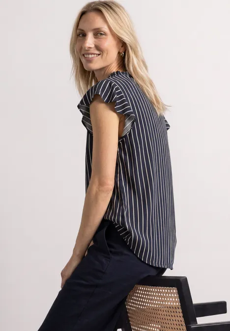 Femme blonde en chemisier rayé assise de profil sur une chaise, souriant à la caméra.