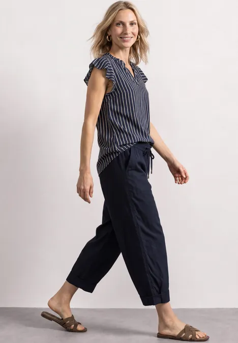 Femme souriante en chemisier rayé, pantalon ample et sandales, marchant légèrement.