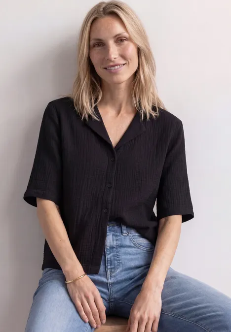 Eine lächelnde Frau mit blonder Mähne, trägt schwarzes Hemd und Jeans, sitzt vor weißer Wand.