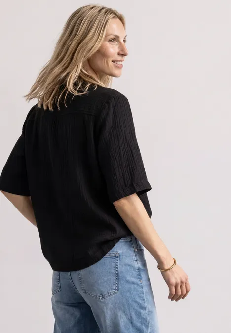 Blonde Frau im Halbprofil mit schwarzem, lockerem Oberteil und blauer Jeans vor hellem Hintergrund.