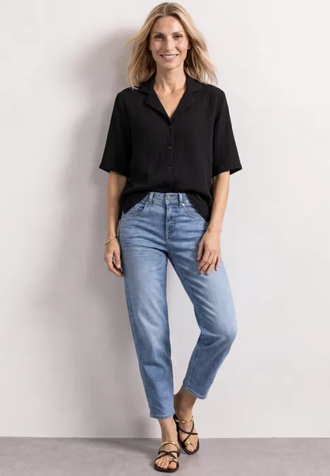 Frau in schwarzer Bluse, Jeans und Sandalen vor neutralem Hintergrund.